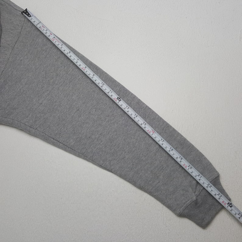 Pode incluir: Uma manga de camisola cinzenta &eacute; medida com uma fita m&eacute;trica amarela. A manga est&aacute; colocada sobre uma superf&iacute;cie branca. A fita m&eacute;trica mostra um comprimento de aproximadamente 61 cm.