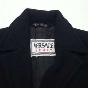 Może przedstawiać: Czarny płaszcz Versace Sport z wykładanym kołnierzem. Płaszcz posiada etykietę z napisem "Versace Sport" w białym prostokącie z czerwonymi akcentami. Podszewka jest czarna i pikowana. Etykieta głosi "Made in Italy".