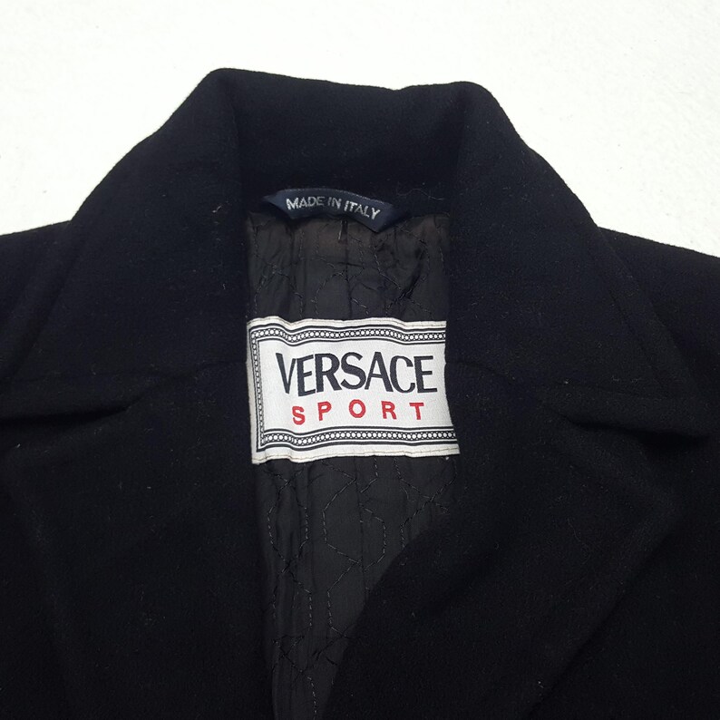 Może przedstawiać: Czarny płaszcz Versace Sport z wykładanym kołnierzem. Płaszcz posiada etykietę z napisem "Versace Sport" w białym prostokącie z czerwonymi akcentami. Podszewka jest czarna i pikowana. Etykieta głosi "Made in Italy".