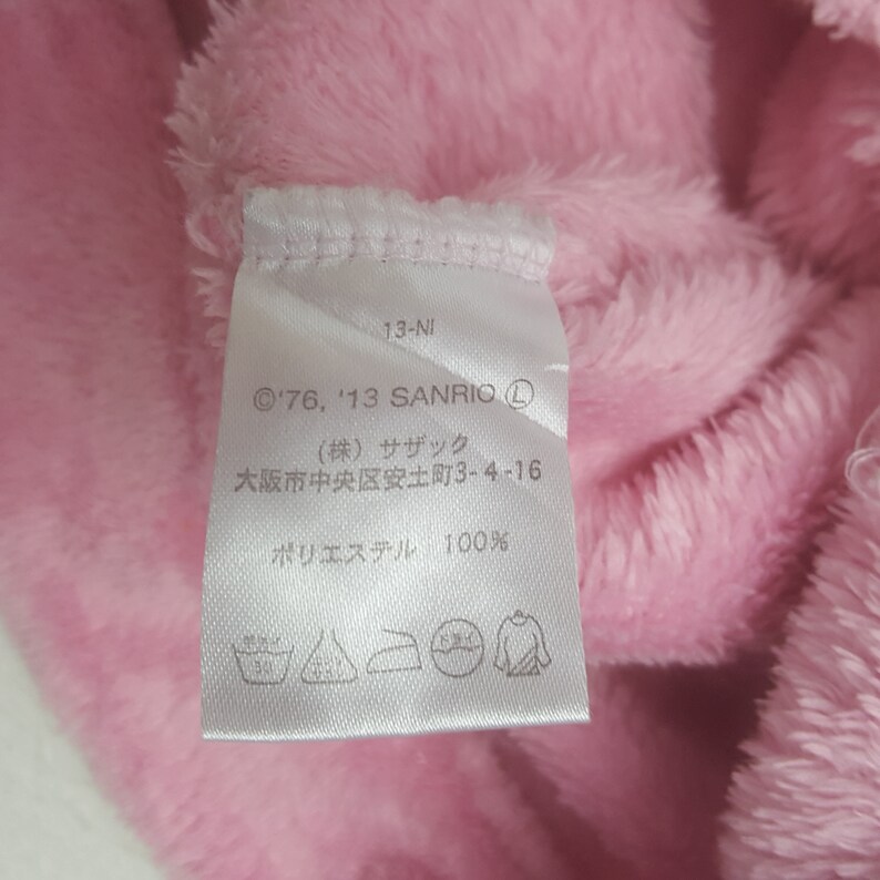 May include: Close-up of a pink, fluffy fabric with a white tag attached. The tag has text including "'76, '13 SANRIO" and Japanese characters. The fabric appears to be made of polyester.