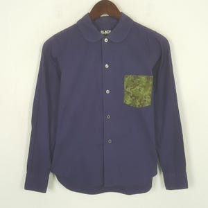 Puede incluir: Una camisa azul marino de manga larga con cuello redondo y un bolsillo en el pecho. El bolsillo presenta un estampado de camuflaje en tonos verdes, marrones y beige. La camisa tiene un diseño clásico con botones.