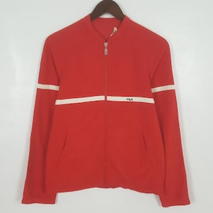 May include: Red zip-up jacket with white stripes on the sleeves and chest. The jacket has a stand-up collar and a small "FILA" logo on the chest. The jacket is hanging on a wooden hanger.
