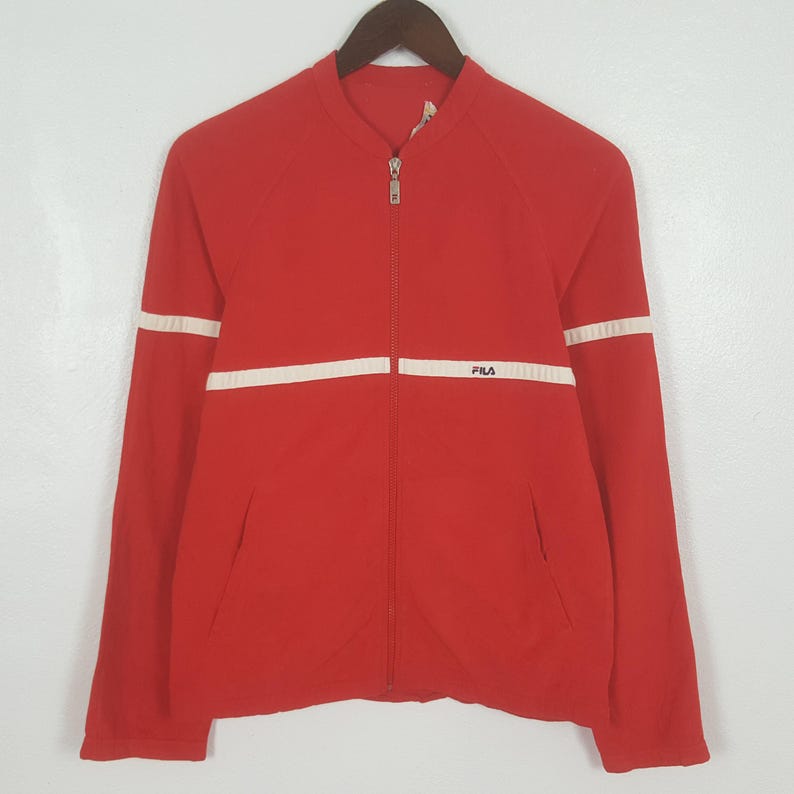 May include: Red zip-up jacket with white stripes on the sleeves and chest. The jacket has a stand-up collar and a small "FILA" logo on the chest. The jacket is hanging on a wooden hanger.
