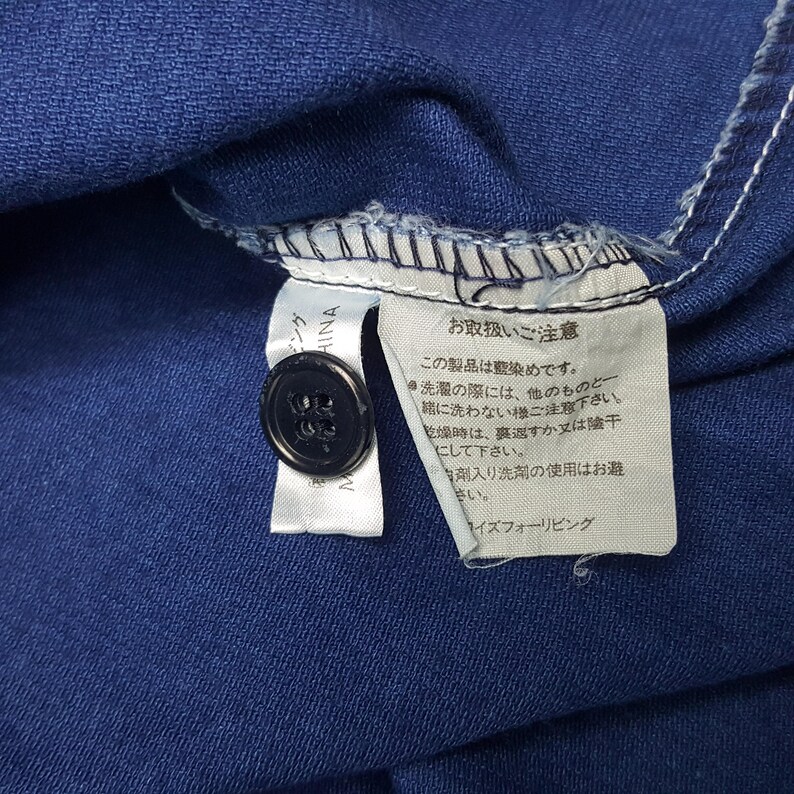 May include: Close-up of a navy blue garment with a black button and a white tag. The tag has Japanese text and care instructions. The garment is made in China.