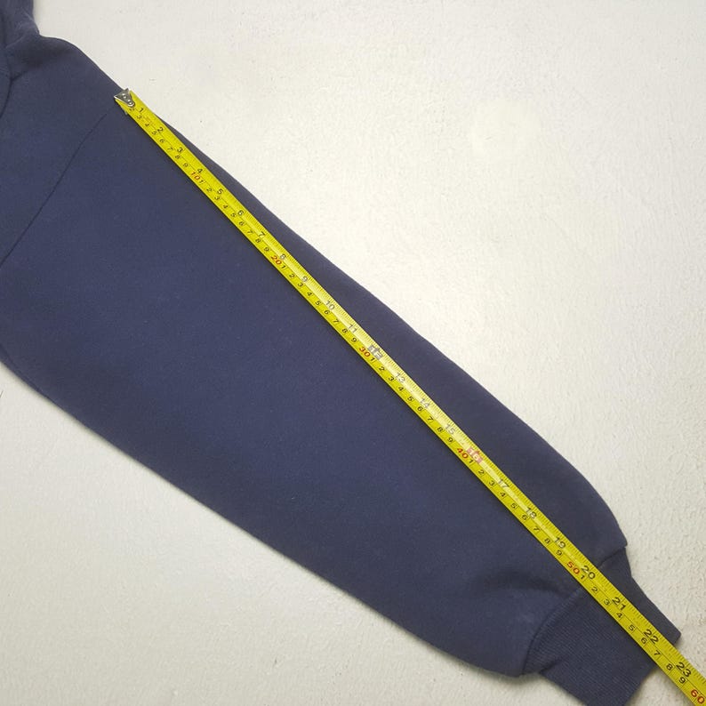 Puede incluir: Una manga de sudadera azul marino se mide con una cinta m&eacute;trica amarilla. La manga est&aacute; colocada sobre una superficie blanca. La cinta m&eacute;trica muestra una longitud de aproximadamente 58 cm.
