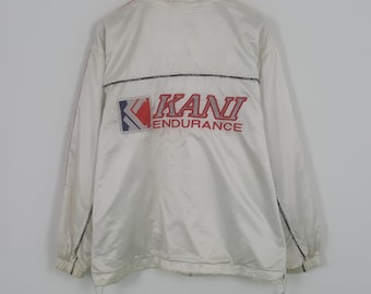 May include: White Kani windbreaker jacket with red and blue logo details. The jacket features the Kani logo on the collar and back, with "KANI ENDURANCE" printed on the back. The jacket has a full zip and drawstring at the bottom.