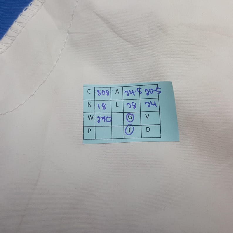 Puede incluir: Primer plano de una tela blanca con una etiqueta azul. La etiqueta tiene texto escrito a mano en tinta azul, incluyendo n&uacute;meros y letras. La tela parece lisa, posiblemente parte de una prenda.