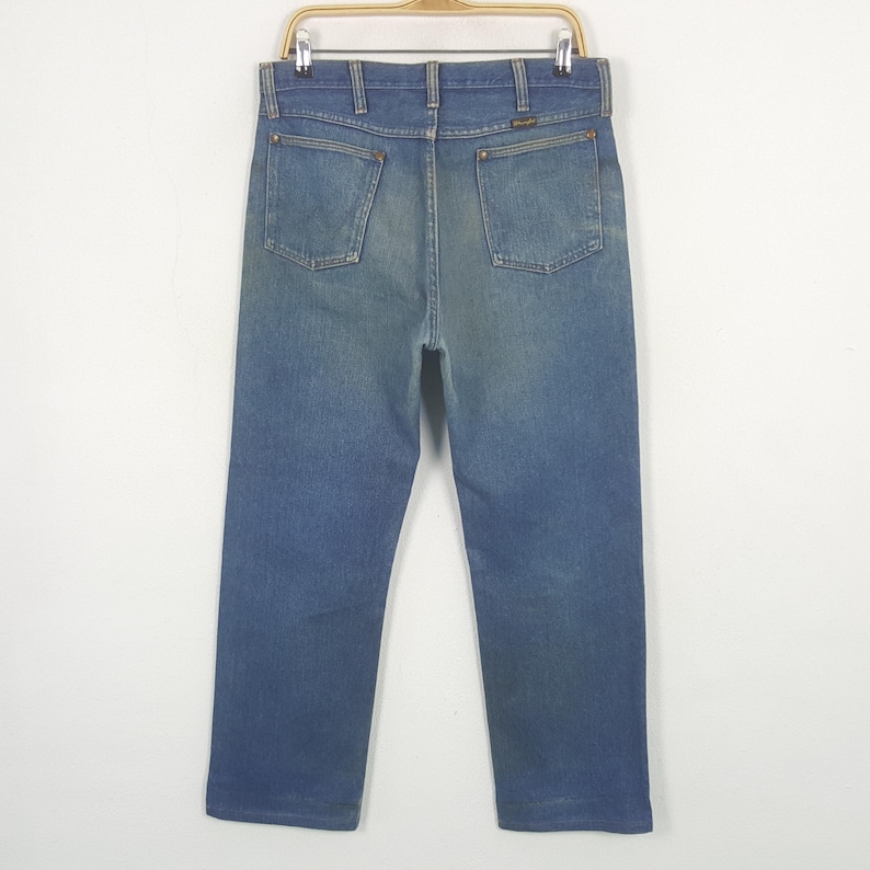 May include: Blue denim jeans with a classic five-pocket design, hanging on a metal hanger. The jeans have a faded wash and a straight leg cut. The Wrangler logo is visible on the back pocket. The jeans are a medium wash blue colour.