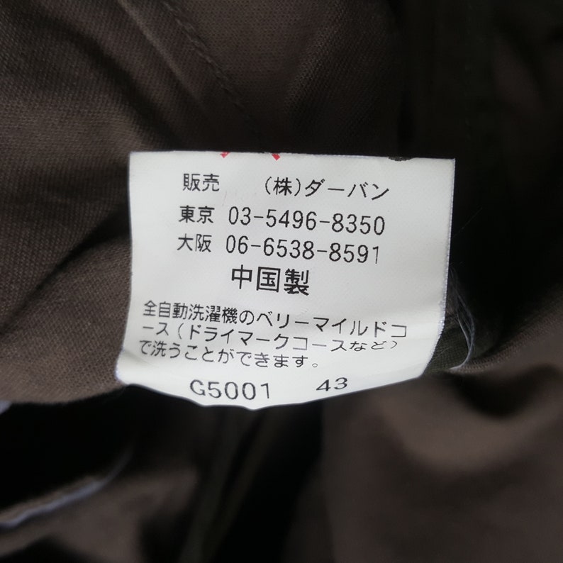 Puede incluir: Primer plano de una etiqueta blanca con texto y n&uacute;meros japoneses. La etiqueta est&aacute; adherida a una tela marr&oacute;n. El texto incluye el nombre de la marca, n&uacute;meros de tel&eacute;fono e instrucciones de lavado. La etiqueta tambi&eacute;n tiene los n&uacute;meros "G5001" y "43".