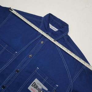 May include: A dark blue button-down shirt with white stitching and two front pockets. The shirt has a collar and the text "YS FOR LIVING" is visible on the collar. A measuring tape is laid across the shirt.