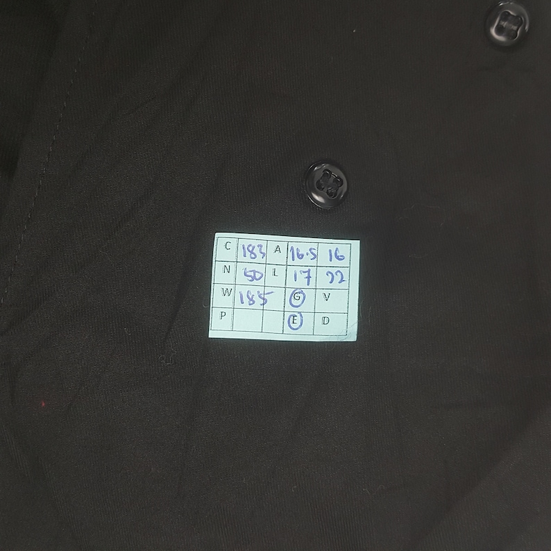 May include: A black button-down shirt with two visible black buttons. A small, rectangular tag with handwritten measurements is attached to the shirt. The shirt's fabric appears smooth and opaque.