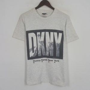 May include: Pale grey DKNY t-shirt with a black rectangular graphic. The graphic features the brand name, a grayscale image of the Statue of Liberty and New York City, and the text "Donna Karan New York".