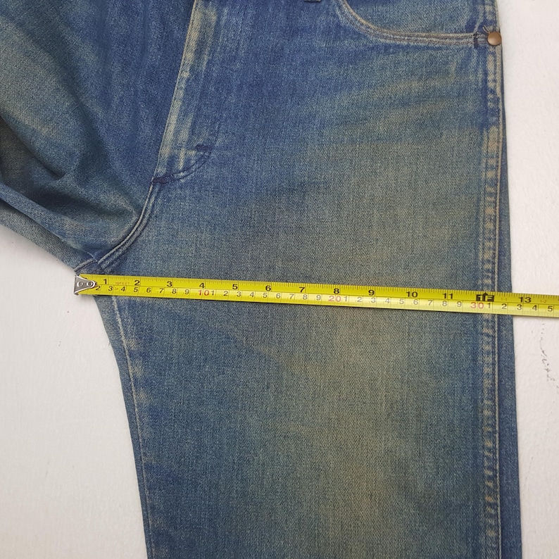 May include: Close-up of blue denim jeans with a yellow measuring tape showing a measurement of approximately 33 cm. The jeans have a faded appearance and are laid flat on a white surface.