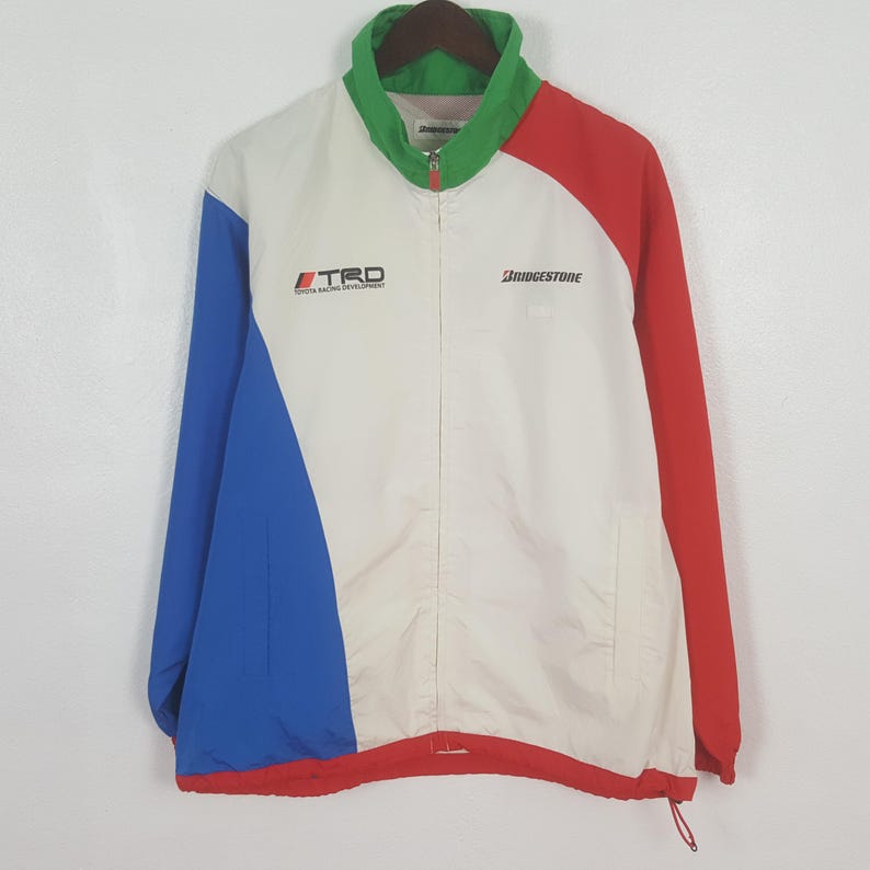 Puede incluir: Chaqueta de carreras vintage con dise&ntilde;o de bloques de color. La chaqueta presenta un cuerpo blanco con detalles en rojo, azul y verde. Los logotipos de "TRD Toyota Racing Development" y "Bridgestone" son visibles en la parte delantera.