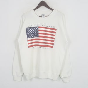 May include: A white sweatshirt featuring a graphic of the American flag. The flag design includes red and white stripes and a blue field with white stars. The text "UNITED STATES OF AMERICA" is printed above and below the flag.