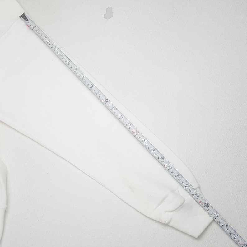May include: A white long-sleeved sweatshirt with a tape measure laid across the sleeve. The tape measure shows measurements in inches, with the sleeve measuring approximately 28 inches in length. The sweatshirt is on a white background.