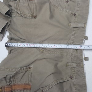May include: Khaki-colored cargo shorts with multiple pockets and brown leather accents. The shorts are measured with a tape measure, showing a length of approximately 12 inches.