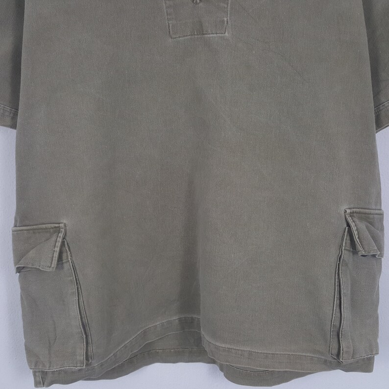 Puede incluir: Camiseta verde oliva de manga corta con dos bolsillos delanteros y un peque&ntilde;o cuello. La camisa tiene un corte relajado y est&aacute; hecha de una tela duradera. La camisa es de color liso y tiene un estilo casual.