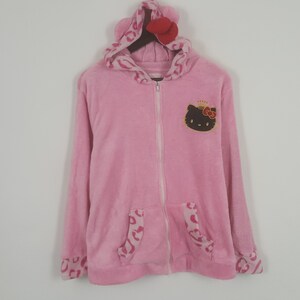 May include: Pink zip-up hoodie with a Hello Kitty graphic on the chest. The hoodie features a hood with ears and a red bow, along with leopard print accents on the pockets and cuffs. The plush material gives a soft appearance.