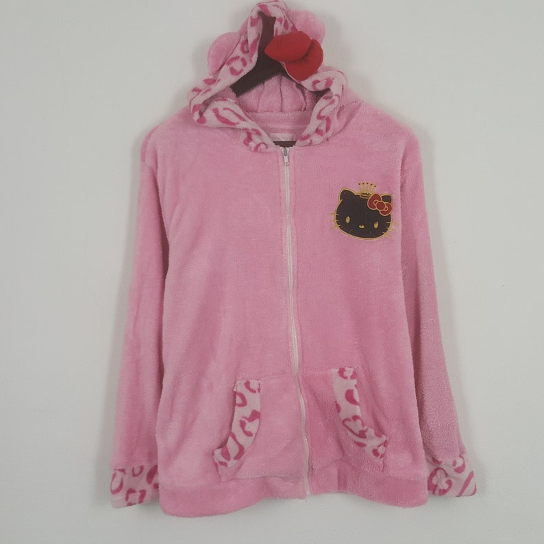 May include: Pink zip-up hoodie with a Hello Kitty graphic on the chest. The hoodie features a hood with ears and a red bow, along with leopard print accents on the pockets and cuffs. The plush material gives a soft appearance.