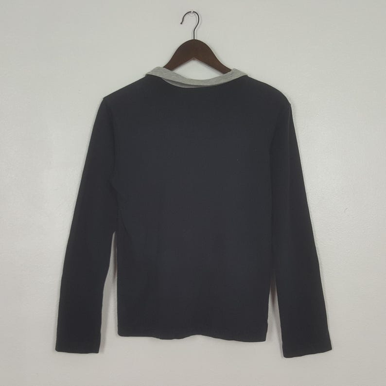 K&ouml;nnte beinhalten: Ein schwarzes Langarmshirt mit grauem Kragen, das an einem Holzkleiderb&uuml;gel h&auml;ngt. Das Shirt besteht aus einem einfarbigen schwarzen Material. Der Kragen ist ein hellerer Grauton.