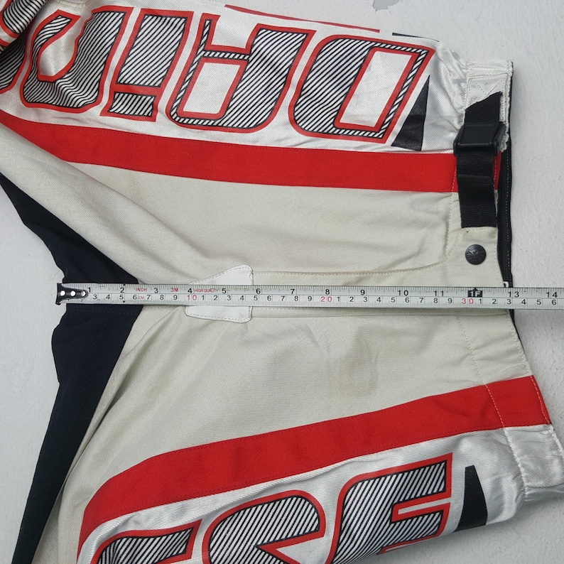 Peut inclure: Pantalon de motocross blanc et cr&egrave;me avec des d&eacute;tails rouges et noirs. Le mot "DARE" est imprim&eacute; en gros caract&egrave;res ray&eacute;s. Un m&egrave;tre ruban indique une longueur d'environ 33 cm.