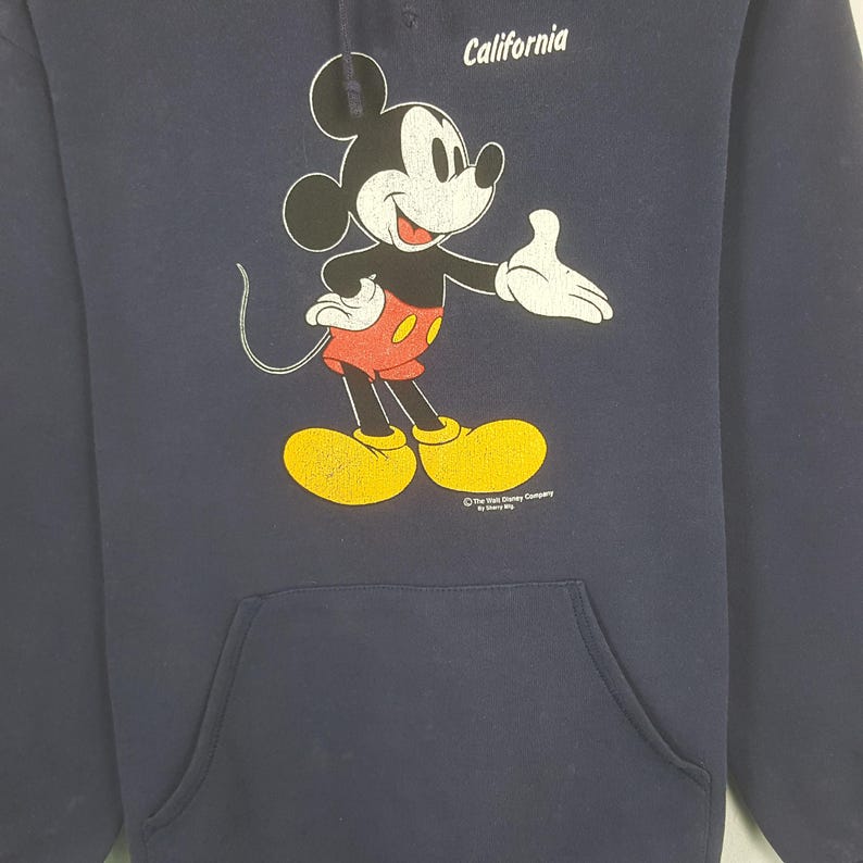 Puede incluir: Sudadera con capucha azul marino con un dise&ntilde;o de Mickey Mouse. Mickey lleva pantalones cortos rojos y zapatos amarillos. La palabra "California" est&aacute; impresa encima del dise&ntilde;o. El logotipo de The Walt Disney Company est&aacute; en la parte inferior.