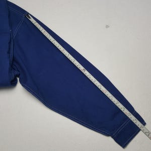 May include: A close-up of a blue long-sleeve shirt with white stitching. A measuring tape is laid across the sleeve, showing a length of approximately 23 inches. The shirt is made of a solid blue fabric.