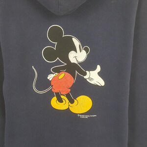 Puede incluir: Sudadera con capucha azul marino con un gr&aacute;fico de Mickey Mouse. El personaje de dibujos animados est&aacute; en negro, blanco, rojo y amarillo. La sudadera tiene una capucha con cord&oacute;n y un bolsillo de canguro. Logotipo de The Walt Disney Company.