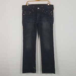 May include: Dark wash denim jeans with a classic five-pocket design. The jeans are hung on a wooden hanger against a white background. Features include a button closure, belt loops, and subtle distressing.