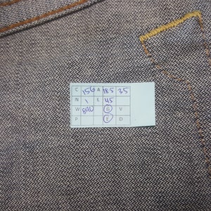 May include: Close-up of denim fabric with visible stitching and a small white tag with handwritten numbers and letters. The denim has a textured appearance with a mix of dark and light threads. The stitching is in a contrasting tan colour.