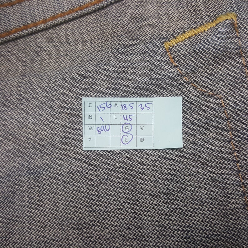 May include: Close-up of denim fabric with visible stitching and a small white tag with handwritten numbers and letters. The denim has a textured appearance with a mix of dark and light threads. The stitching is in a contrasting tan colour.
