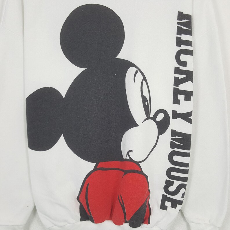 Może przedstawiać: Biała bluza z dużą czarno-czerwoną grafiką Myszki Miki. Słowa "MICKEY MOUSE" są wydrukowane pionowo w kolorze czarnym na prawym rękawie. Projekt jest w klasycznym stylu kresk&oacute;wkowym.