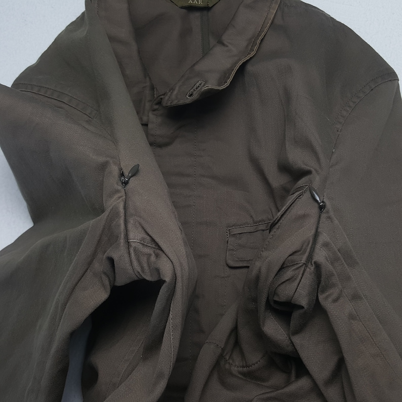Puede incluir: Chaqueta verde oliva con cuello abotonado y un peque&ntilde;o bolsillo. La chaqueta tiene dos bolsillos con cremallera en las mangas. La marca "AAR" es visible en el cuello.