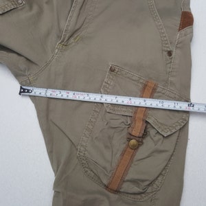 May include: Khaki-colored cargo pants with a brown leather accent on the pocket. The pants are being measured with a tape measure, showing a length of approximately 12 inches. The pants have multiple pockets and a button closure.