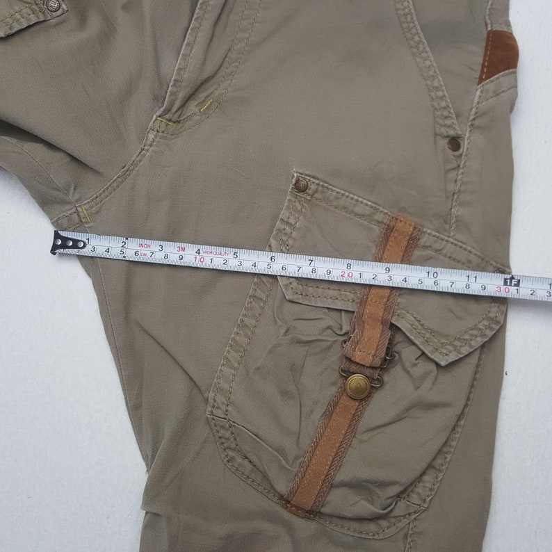 May include: Khaki-colored cargo pants with a brown leather accent on the pocket. The pants are being measured with a tape measure, showing a length of approximately 12 inches. The pants have multiple pockets and a button closure.