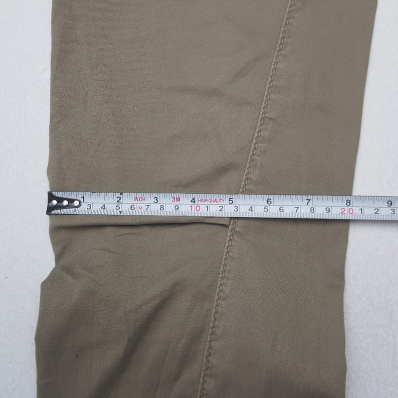 May include: Close-up of tan-colored pants with a measuring tape showing a width of approximately 8 inches. The tape measure is marked in inches and centimeters, indicating the garment's dimensions.