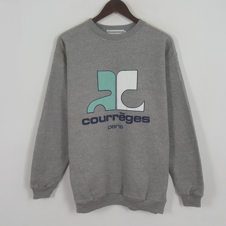 Pode incluir: Um moletom cinza com mangas compridas e gola redonda. A frente apresenta um logotipo com formas turquesa e brancas, e o texto "courr&egrave;ges paris" em azul marinho. O moletom est&aacute; pendurado em um cabide de madeira.