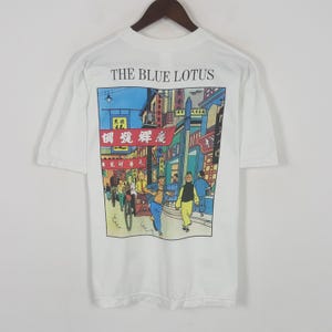 May include: White t-shirt featuring a colorful illustration from "The Blue Lotus" comic. The graphic depicts a bustling street scene with figures, buildings, and text in a foreign language. The words "THE BLUE LOTUS" are printed above the illustration.