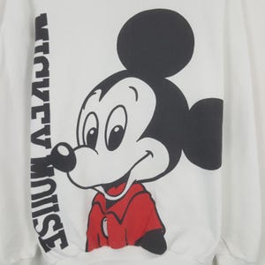 Może przedstawiać: Biała bluza z dużym nadrukiem Myszki Miki w kolorach czarnym, czerwonym i białym. Tekst "MICKEY MOUSE" jest wydrukowany pionowo po lewej stronie. Projekt jest w klasycznym stylu kresk&oacute;wki.