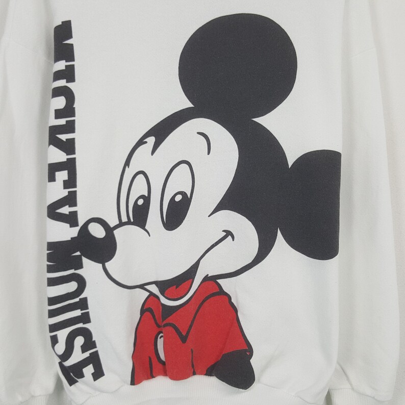 Może przedstawiać: Biała bluza z dużym nadrukiem Myszki Miki w kolorach czarnym, czerwonym i białym. Tekst "MICKEY MOUSE" jest wydrukowany pionowo po lewej stronie. Projekt jest w klasycznym stylu kresk&oacute;wki.