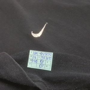 Peut inclure: V&ecirc;tement noir avec un logo Nike swoosh blanc brod&eacute;. Une petite &eacute;tiquette manuscrite avec des chiffres et des lettres est visible. Le tissu semble &ecirc;tre doux, peut-&ecirc;tre du coton ou un m&eacute;lange.