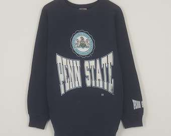 Könnte beinhalten: Marineblaues Penn State University Sweatshirt. Auf der Vorderseite stehen die Worte "PENN STATE" in weißen Blockbuchstaben, darüber ein Universitätslogo. Auf dem Ärmel steht "PENN STATE".