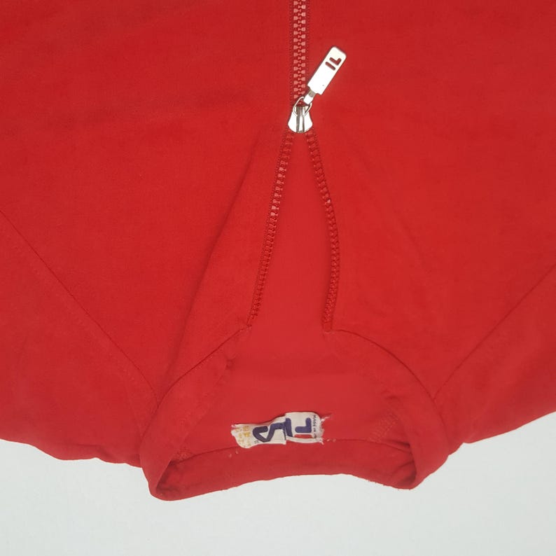 May include: Close-up of a bright red athletic jacket with a silver zipper and the brand logo. The zipper is partially open, revealing the fabric underneath. The jacket has a sporty, casual style.