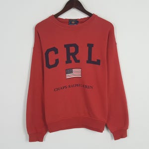 Puede incluir: Sudadera roja con las letras "CRL" en azul marino en el pecho, una pequeña bandera estadounidense y la inscripción "CHAPS-RALPH LAUREN". La sudadera está colgada de una percha de madera.