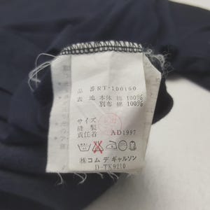 K&ouml;nnte beinhalten: Nahaufnahme eines wei&szlig;en Kleidungsetiketts mit japanischem Text und Symbolen. Das Etikett ist an einem marineblauen Kleidungsst&uuml;ck befestigt. Das Etikett enth&auml;lt Waschanweisungen und den Text "RT-100160".