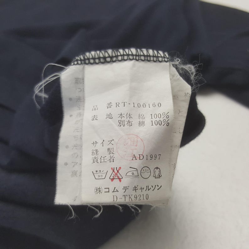 K&ouml;nnte beinhalten: Nahaufnahme eines wei&szlig;en Kleidungsetiketts mit japanischem Text und Symbolen. Das Etikett ist an einem marineblauen Kleidungsst&uuml;ck befestigt. Das Etikett enth&auml;lt Waschanweisungen und den Text "RT-100160".