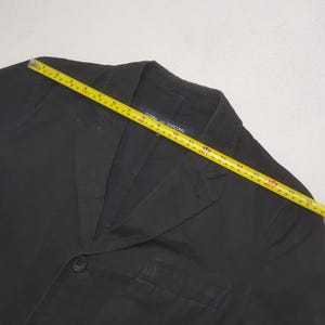 Puede incluir: Blazer negro con cierre de un solo bot&oacute;n y solapas con muescas. Una cinta m&eacute;trica amarilla est&aacute; colocada sobre el blazer. La etiqueta dice "Comme des Garcons".