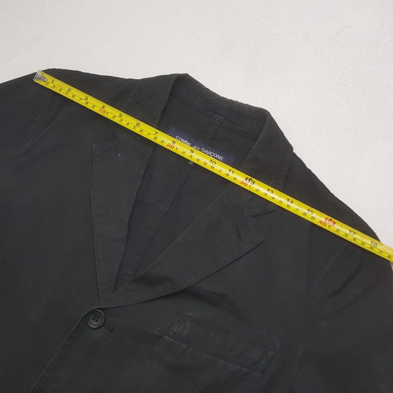 Puede incluir: Blazer negro con cierre de un solo bot&oacute;n y solapas con muescas. Una cinta m&eacute;trica amarilla est&aacute; colocada sobre el blazer. La etiqueta dice "Comme des Garcons".