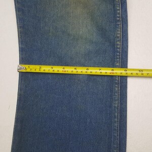 May include: Close-up of blue denim fabric with a yellow measuring tape. The tape shows measurements in centimetres, highlighting the width of the denim. The fabric has a faded appearance, with visible seams.